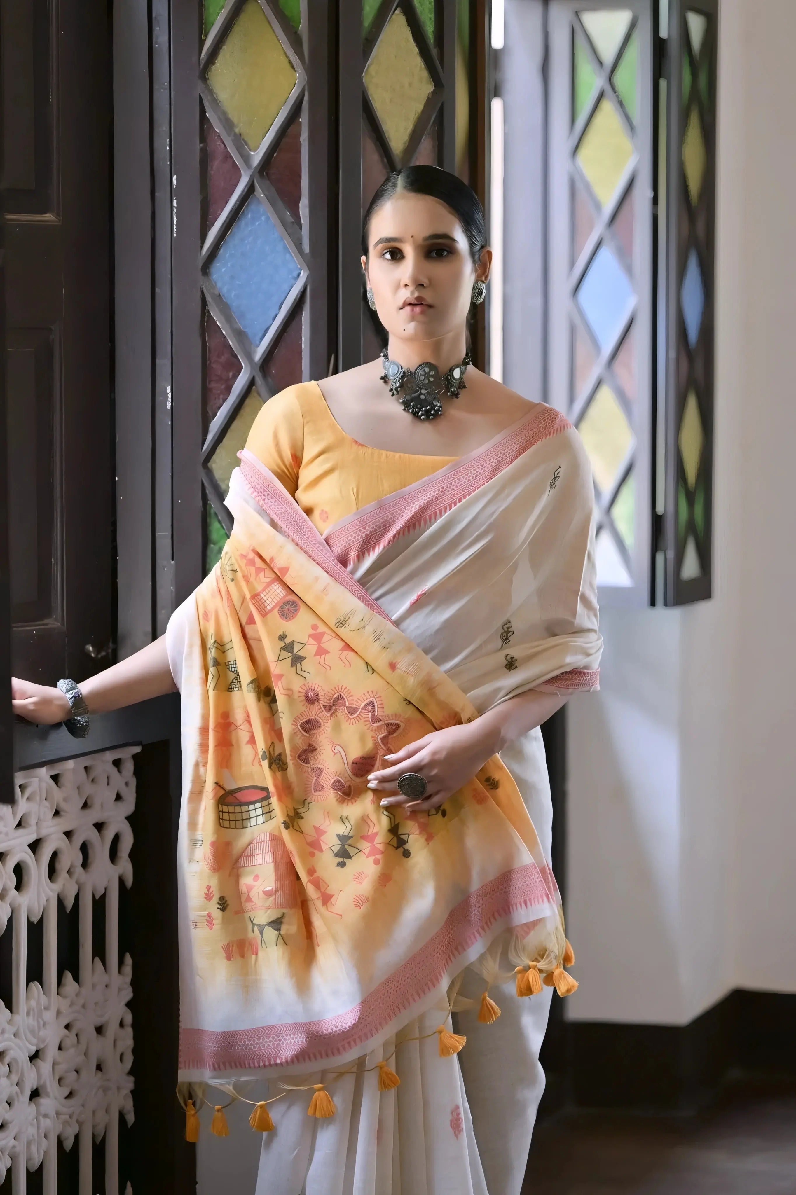 A woman models a graceful Off White Soft Muga Cotton Silk Saree. The saree has a pink accent border, and the pallu features a wide yellow panel decorated with traditional Warli-style figurative motifs. She wears a yellow blouse and a dark oxidized choker necklace, posing indoors.